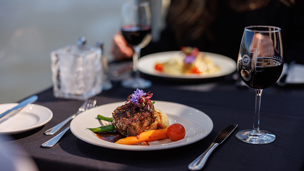 A gourmet dish is served on an elegant table covered with a dark tablecloth. The main course, garnished with vegetables and edible flowers, is accompanied by glasses of red wine. In the background, another plate is visible, enhancing the refined dining atmosphere.