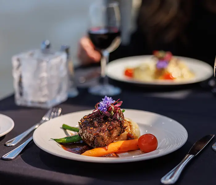Un plat gastronomique est servi sur une table élégante recouverte d'une nappe sombre. Le plat principal, décoré de légumes et de fleurs comestibles, est accompagné de verres de vin rouge. À l'arrière-plan, une autre assiette est visible, ajoutant à l'ambiance raffinée du repas.