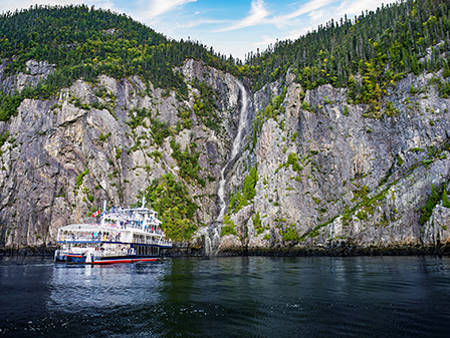 A cruise ship is docked near a towering rocky cliff covered in vegetation. A narrow waterfall flows down the cliff, cascading into the water below, under a blue sky with a few clouds.