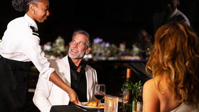 A well-dressed couple dining at a romantic table, served by a smiling waitress. The table is adorned with candles, wine, and plates of fine dishes, with a blurred nighttime view in the background.