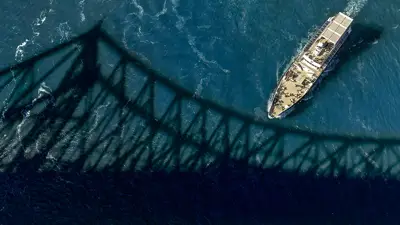 Aerial view of a boat sailing under a bridge, with the bridge's shadow cast on the blue surface of the water.