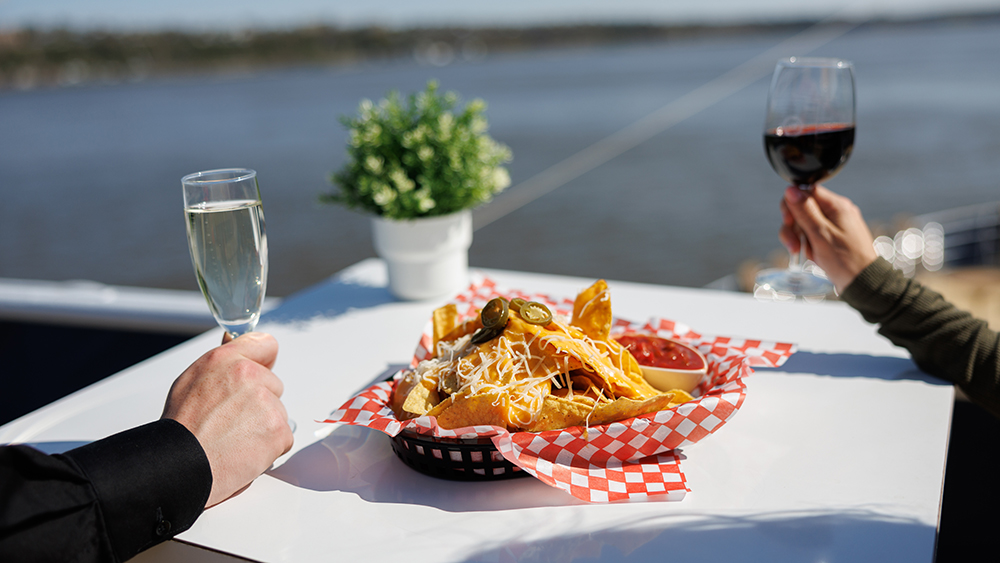 A white table with a basket of nachos topped with cheese and jalapeños, served with red sauce. One person holds a glass of red wine, and another a glass of white wine, with a river and a plant in the background.