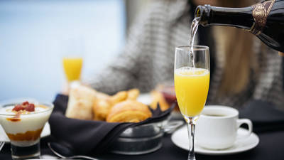 A glass of mimosa is being poured with champagne, accompanied by pastries, a garnished yogurt, and a cup of coffee on an elegant table.
