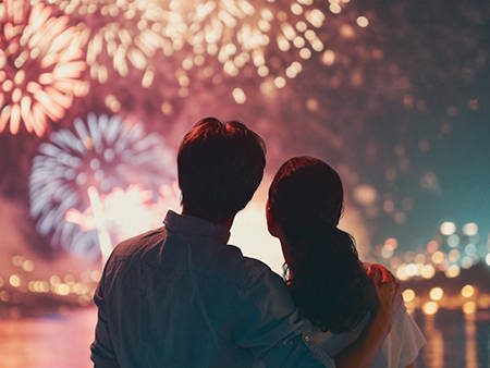 A couple watches a colorful fireworks display lighting up the night sky, with shimmering reflections on the water and city lights in the background.
