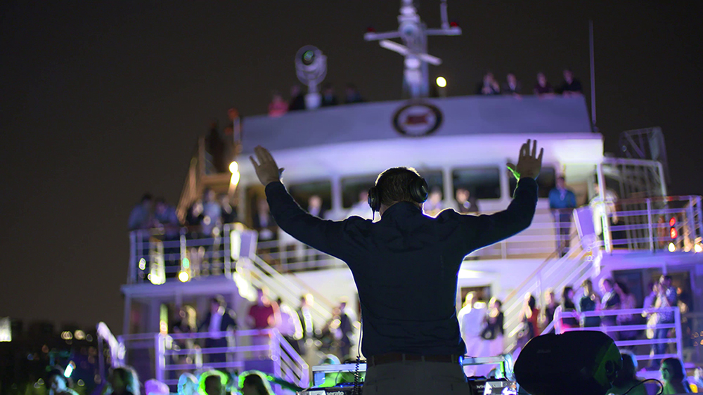 A DJ hosts a lively party on the deck of a boat, arms raised, with a crowd of attendees and colorful lights illuminating the scene at night.