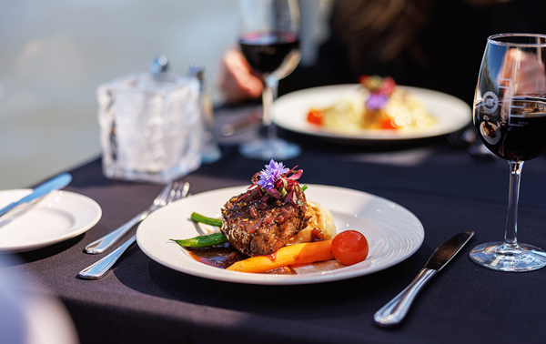A gourmet steak dish garnished with a flower, paired with a glass of red wine, overlooking the serene backdrop of a water view.