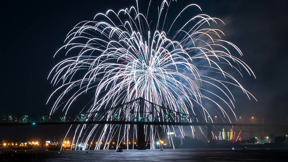 Un spectaculaire feu d'artifice illumine le ciel nocturne au-dessus du pont Jacques-Cartier, avec ses reflets brillant sur le fleuve.