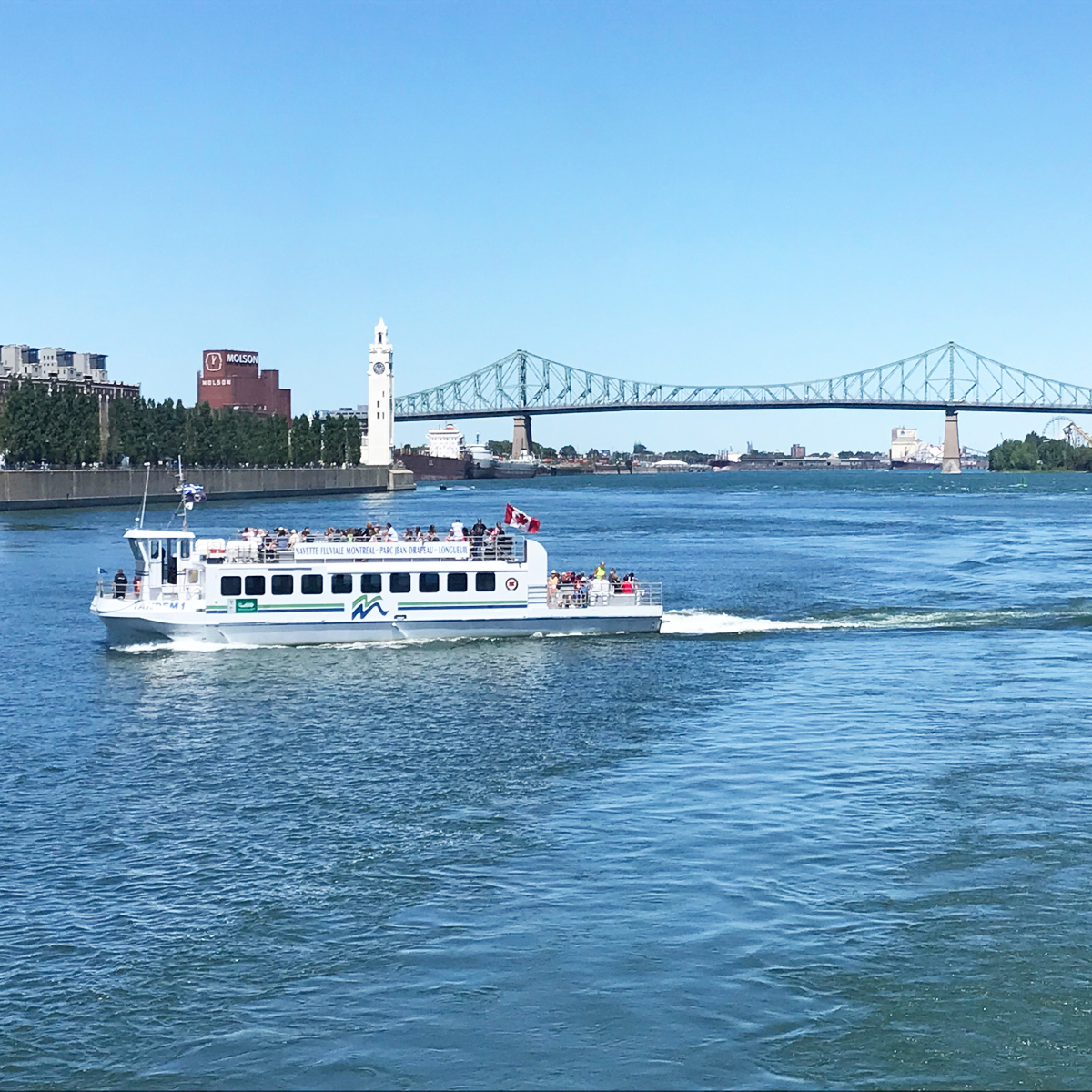 Une navette fluviale traverse le fleuve sous un ciel bleu, avec la tour de l'Horloge, le pont Jacques-Cartier et des bâtiments de Montréal visibles en arrière-plan.
