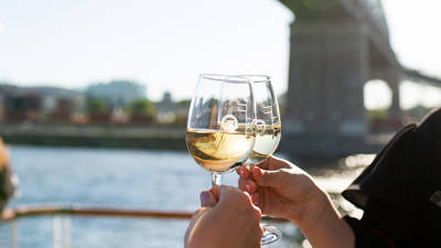 Two people clinking glasses of white wine on an outdoor terrace, with a river, a bridge, and buildings in the background on a sunny day.