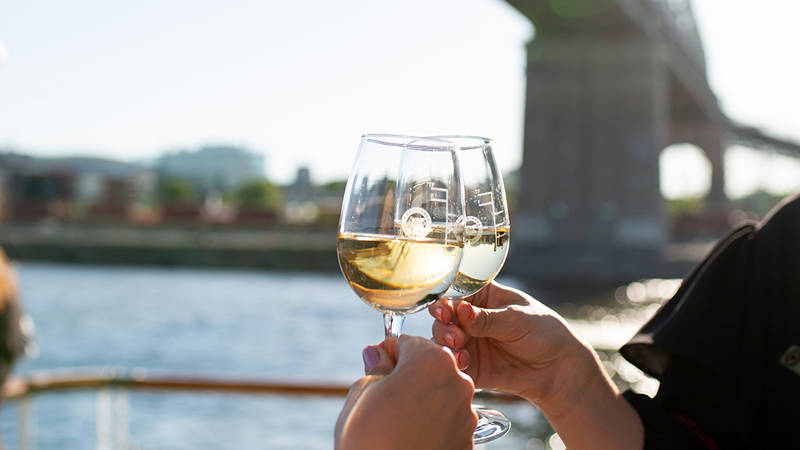 Two people clinking glasses of white wine on an outdoor terrace, with a river, a bridge, and buildings in the background on a sunny day.
