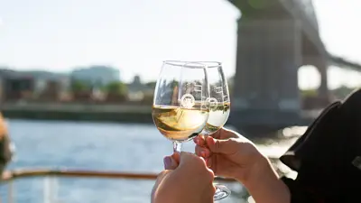 Two people clinking glasses of white wine on an outdoor terrace, with a river, a bridge, and buildings in the background on a sunny day.