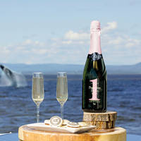  A bottle of sparkling wine and two filled glasses sit on a wooden platter by the ocean. Nearby are gourmet pastries. In the background, a whale breaches from the water under a partly cloudy sky