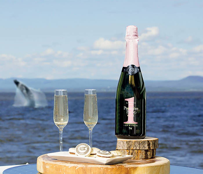 Une bouteille de vin mousseux et deux verres remplis reposent sur un plateau en bois près de l'océan. À proximité, des pâtisseries gourmandes sont disposées. En arrière-plan, une baleine jaillit de l'eau sous un ciel partiellement nuageux.