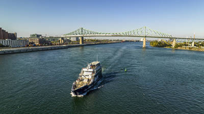 A cruise ship sails on a river beneath a large green bridge, with urban buildings and a clear sky in the background.