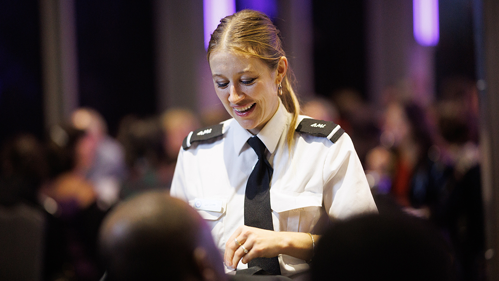 A smiling waitress in professional uniform interacts with customers in an elegant and lively setting, with blurred lights in the background.
