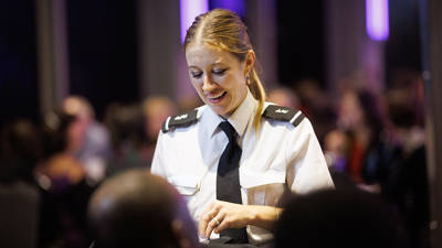A smiling waitress in professional uniform interacts with customers in an elegant and lively setting, with blurred lights in the background.