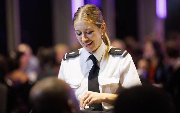 A smiling waitress in professional uniform interacts with customers in an elegant and lively setting, with blurred lights in the background.