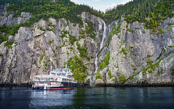A large tour boat sails near towering rocky cliffs with lush green trees, in front of a cascading waterfall that flows into the calm water below. The scene is tranquil, featuring clear skies and reflections on the water's surface.