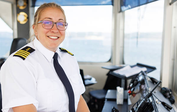 A smiling boat captain, wearing a white uniform with black and gold epaulettes, stands in the ship's cockpit. Through the windows in the background, there is a clear view of the water and sky.
