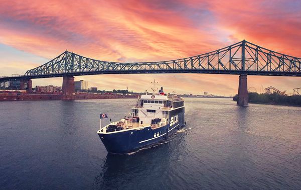 The AML Cavalier Maxim ship sailing alone on the Saint Lawrence River with the Jacques Cartier Bridge and a vibrant sunset in the background.