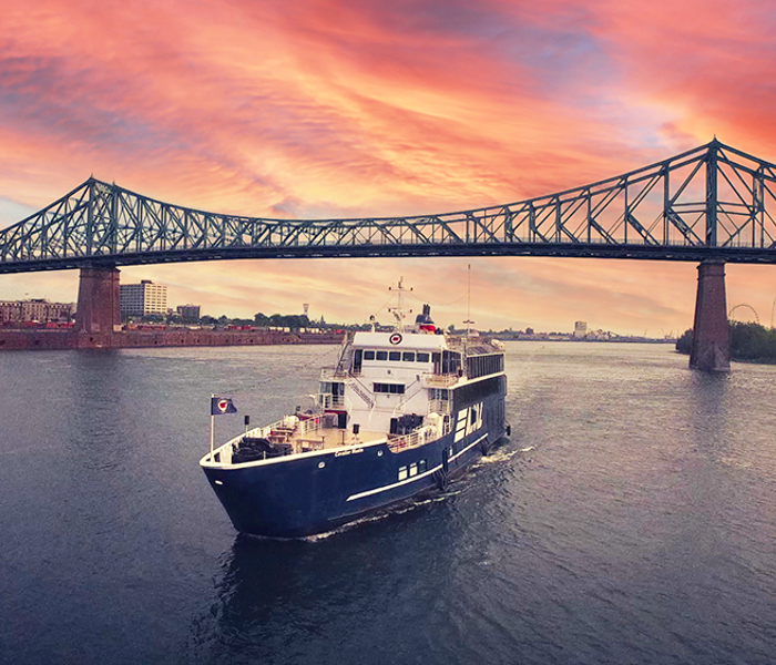 The AML Cavalier Maxim ship sailing alone on the Saint Lawrence River with the Jacques Cartier Bridge and a vibrant sunset in the background.
