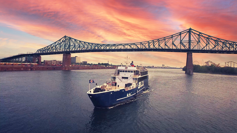 Le navire AML Cavalier Maxim naviguant seul sur le fleuve Saint-Laurent avec le Pont Jacques Cartier et un coucher de soleil vibrant en arrière plan.