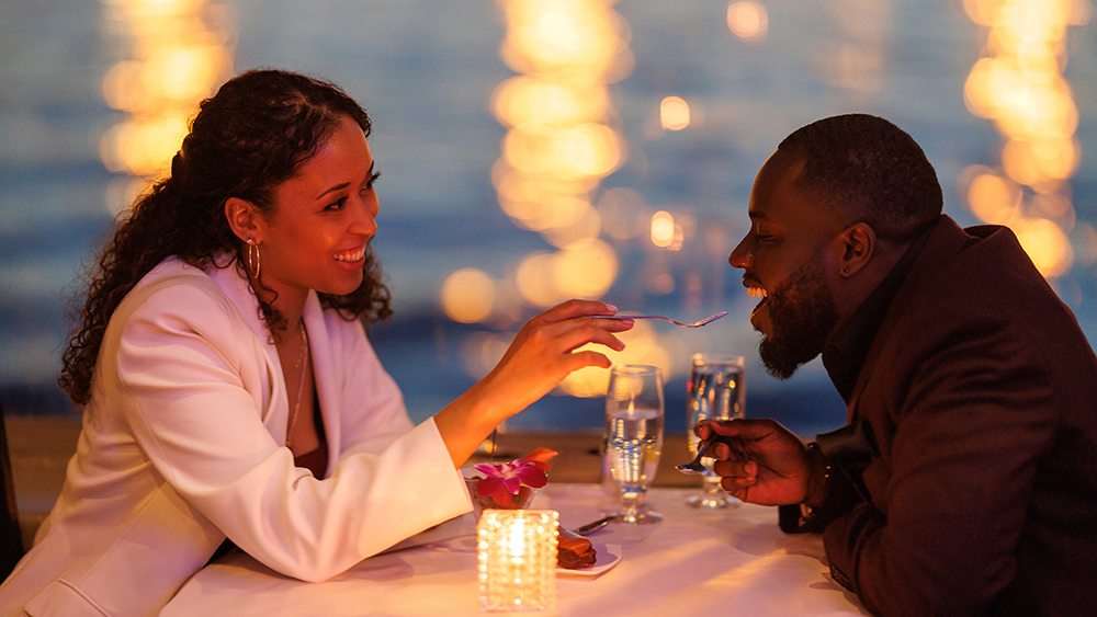 Un couple partage un moment romantique lors d'un dîner aux chandelles près de l'eau. La femme, souriante, donne une bouchée de dessert à l'homme, avec des lumières floues scintillant en arrière-plan.
