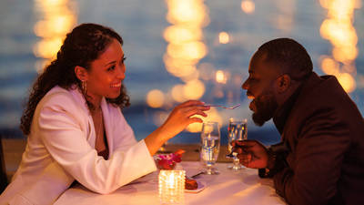 A couple sharing a romantic moment during a candlelit dinner by the water. The smiling woman feeds the man a bite of dessert, with blurred sparkling lights in the background.