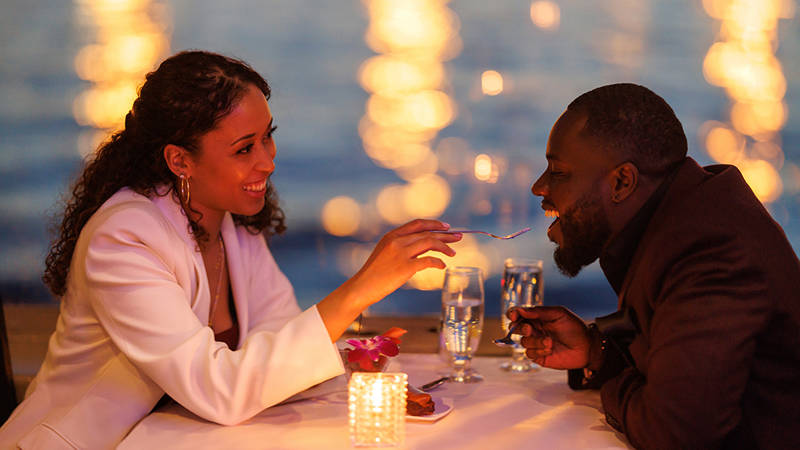 A couple sharing a romantic moment during a candlelit dinner by the water. The smiling woman feeds the man a bite of dessert, with blurred sparkling lights in the background.