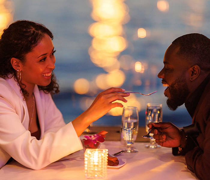 A couple sharing a romantic moment during a candlelit dinner by the water. The smiling woman feeds the man a bite of dessert, with blurred sparkling lights in the background.