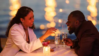 A couple sharing a romantic moment during a candlelit dinner by the water. The smiling woman feeds the man a bite of dessert, with blurred sparkling lights in the background.