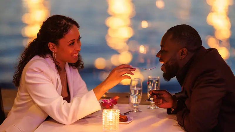 A couple sharing a romantic moment during a candlelit dinner by the water. The smiling woman feeds the man a bite of dessert, with blurred sparkling lights in the background.