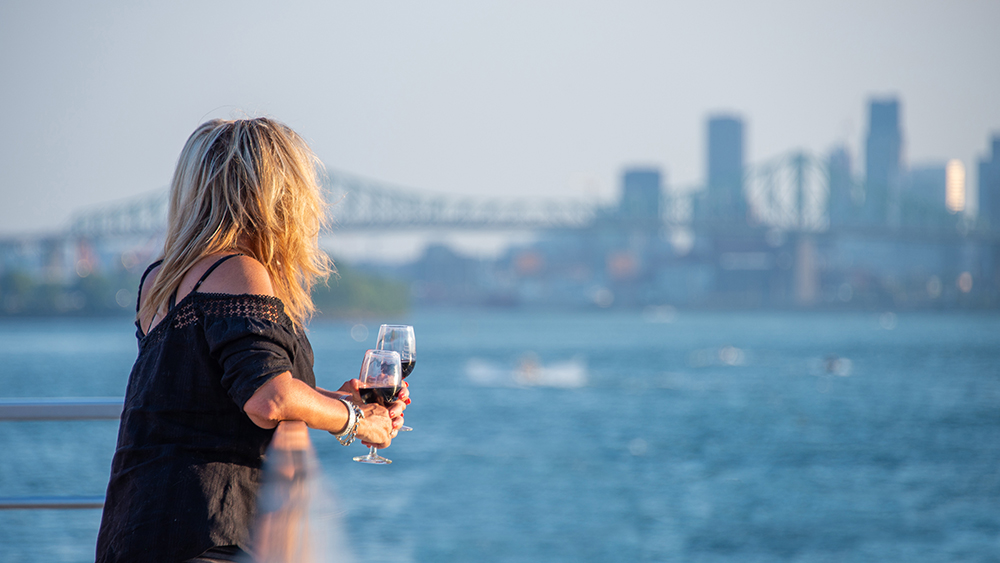 A blonde woman, viewed from behind, holding a glass of red wine, looking at a river with a bridge and skyscrapers in the background on a sunny day.
