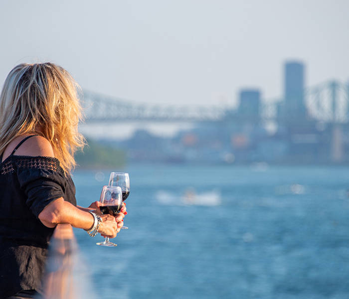 A blonde woman, viewed from behind, holding a glass of red wine, looking at a river with a bridge and skyscrapers in the background on a sunny day.