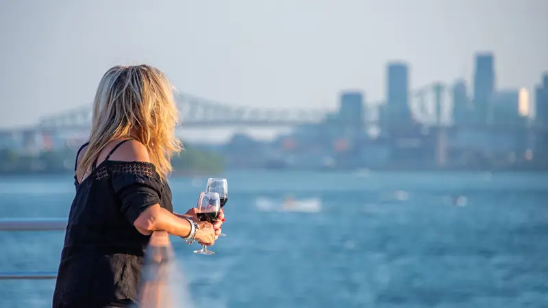 A blonde woman, viewed from behind, holding a glass of red wine, looking at a river with a bridge and skyscrapers in the background on a sunny day.