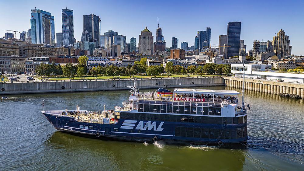 A large black cruise ship with "AML" written on it, sailing on a river. In the background, a Ferris wheel, autumn trees, and urban buildings under a clear blue sky.