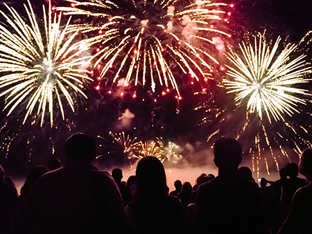Une foule de spectateurs regarde un spectaculaire feu d'artifice illuminant le ciel nocturne avec des éclats dorés et rouges. La scène est empreinte d'émerveillement et de célébration.