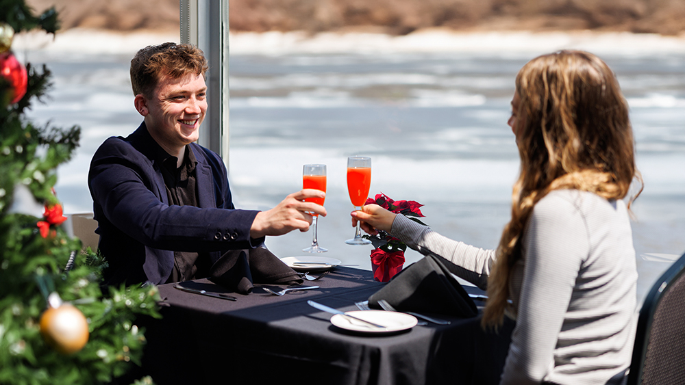 A couple toasts with red cocktails during an elegant meal near a window overlooking a river, with a decorated Christmas tree in the background.