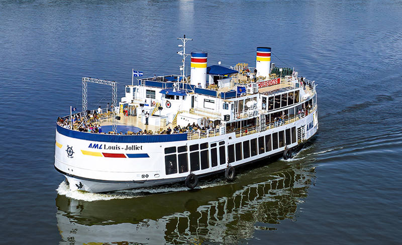 Le bateau "Louis Jolliet" de la compagnie AML navigue sur une rivière calme en pleine journée, avec des passagers sur les ponts observant le paysage environnant. L'eau reflète le bateau et le ciel clair.