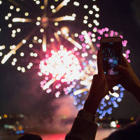 A spectacular firework display bursts in the night sky above an illuminated bridge, with reflections on the water and city lights in the background.