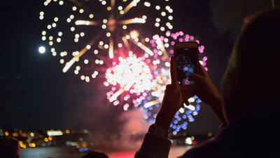 A person holds a smartphone to capture colorful fireworks lighting up the night sky, with reflections on the water and a city in the background.