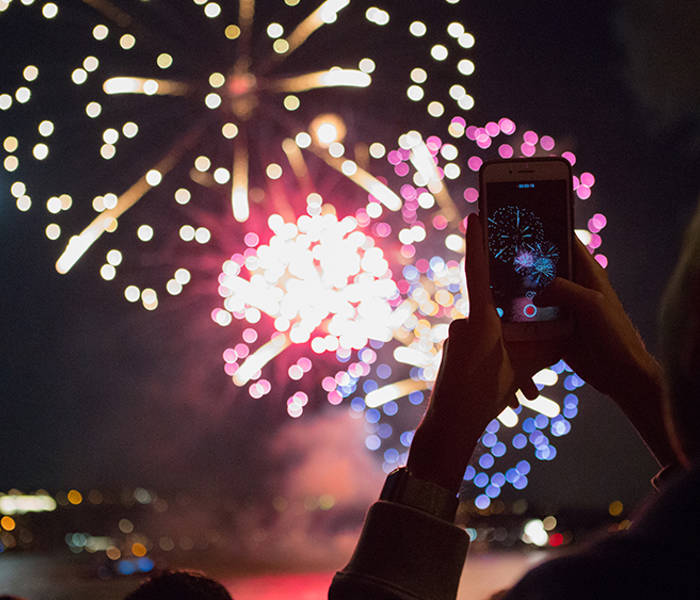 A person holds a smartphone to capture colorful fireworks lighting up the night sky, with reflections on the water and a city in the background.