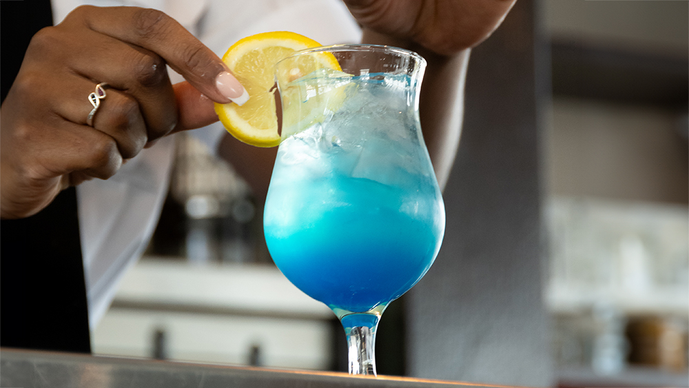 A hand decorates a vibrant blue cocktail with a slice of lemon, adding a finishing touch to the drink served in an elegant glass.