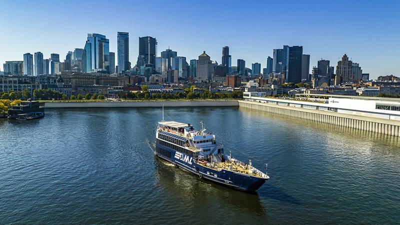 A cruise ship sailing on a river, with a panoramic view of a city skyline featuring tall skyscrapers in the background under a clear sky.
