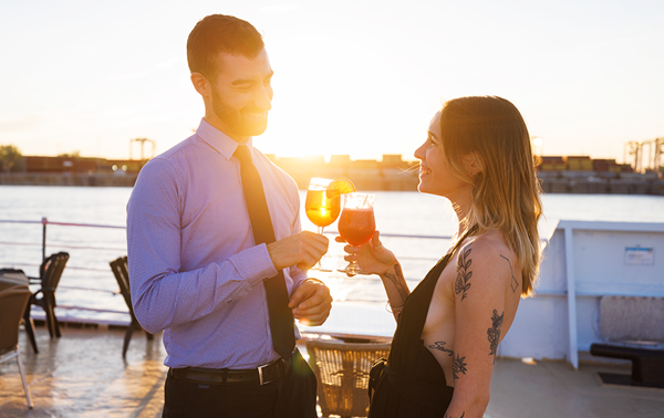 A man and a woman enjoying a drink together on one of the terraces of the AML Cavalier Maxim ship in Montreal, with the sunset in the background.