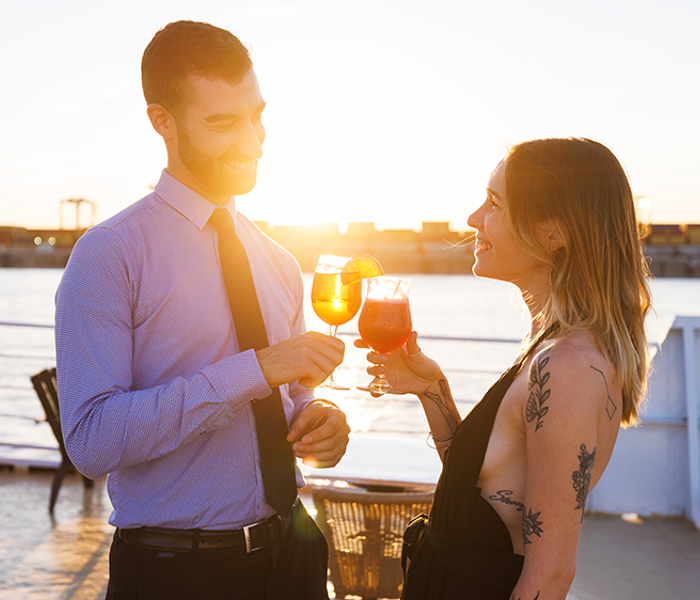 A man and a woman enjoying a drink together on one of the terraces of the AML Cavalier Maxim ship in Montreal, with the sunset in the background.