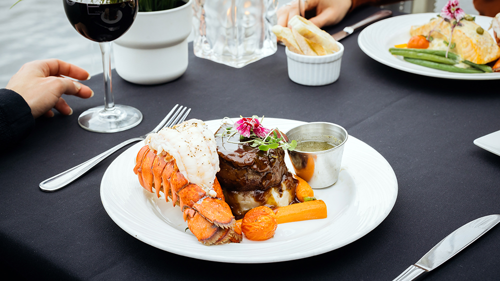A gourmet dish served on a white plate, featuring a lobster tail, a filet of meat with sauce, assorted vegetables, and a floral garnish, placed on an elegant table with a black tablecloth. A glass of red wine is beside it.