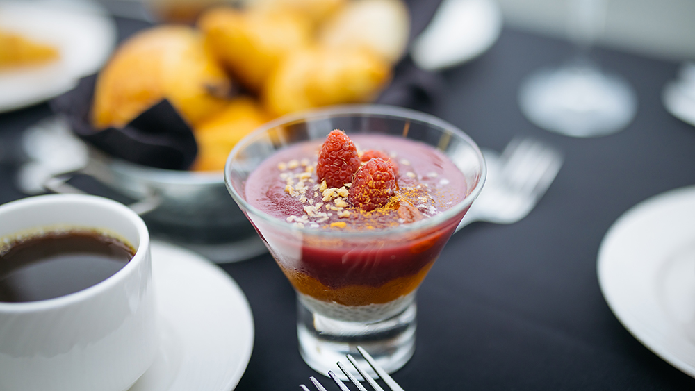 An elegant dessert in a glass, featuring layers of red fruit mousse or puree, topped with fresh raspberries and crumbled nuts or biscuits, accompanied by a cup of coffee and croissants in the background.