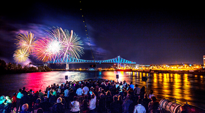 A crowd enjoying a vibrant fireworks display from the main terrace of the AML Cavalier Maxim in Montreal, with a view of the illuminated Jacques Cartier Bridge and city in the background.