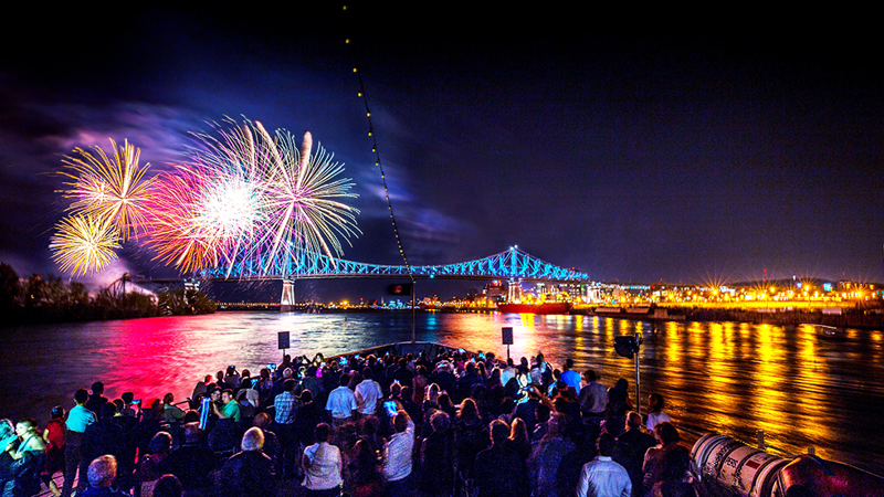 A crowd enjoying a vibrant fireworks display from the main terrace of the AML Cavalier Maxim in Montreal, with a view of the illuminated Jacques Cartier Bridge and city in the background.